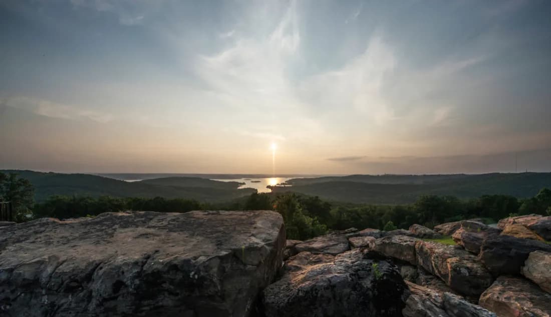 Scenic overlook with rocky foreground and a glowing sunset over Table Rock Lake surrounded by forested hills near Branson, Missouri.