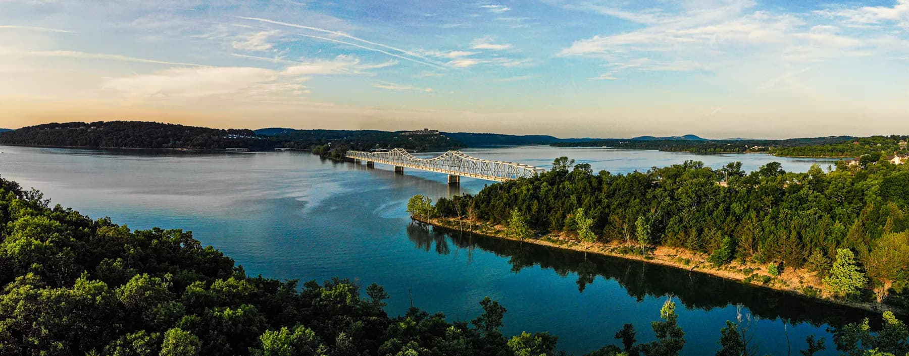 Wide aerial view of Table Rock Lake with a bridge crossing over calm blue water surrounded by green forested hills at sunset.