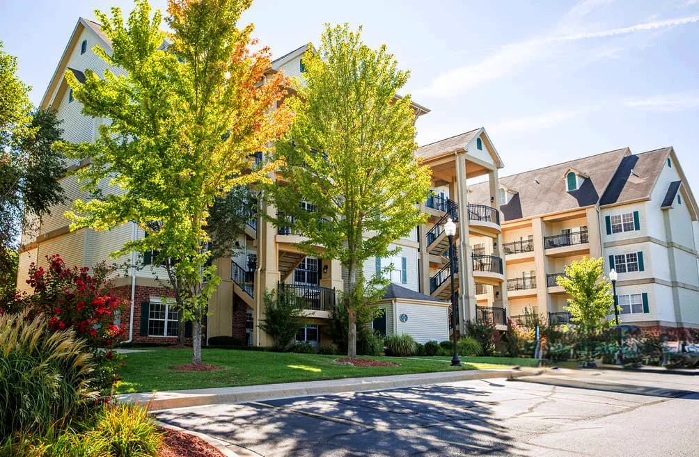 Exterior of a modern multi-story residential building with balconies, tall trees, and landscaped gardens on a bright sunny day.